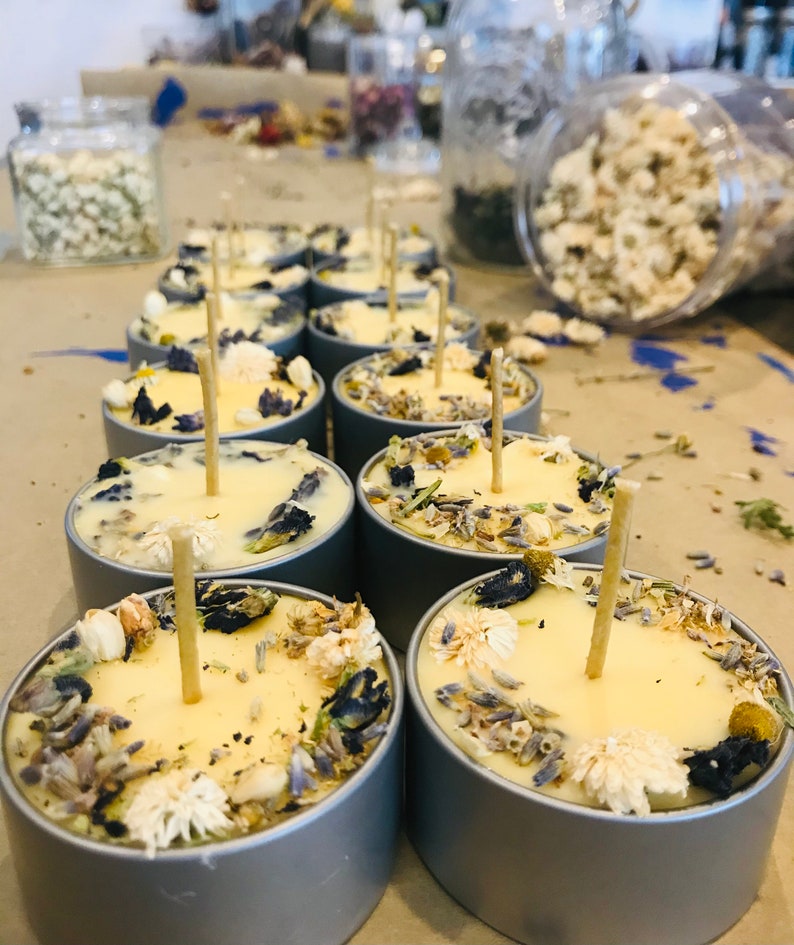 Op de afbeelding: Een close-up van een groep kleine, ronde, gele kaarsen met gedroogde bloemen en kruiden die in de was zijn ingebed. De kaarsen bevinden zich in zilveren containers en hebben houten lonten.