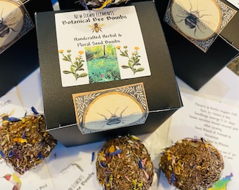 May include: Several botanical bee bombs are displayed, some in black boxes. The bombs are round and covered in dried flowers and seeds. The boxes have labels with a bee illustration and the text "New Dawn Elements Botanical Bee Bombs."