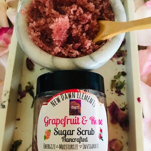 May include: A glass jar of pink grapefruit and rose sugar scrub with a black lid. The label reads "NEW DAWN ELEMENTS Grapefruit & Rose Sugar Scrub Hancrafted ENERGIZE MOISTURIZE INVIGORATE All Natural Skin Care Nt Wt. 4 oz".