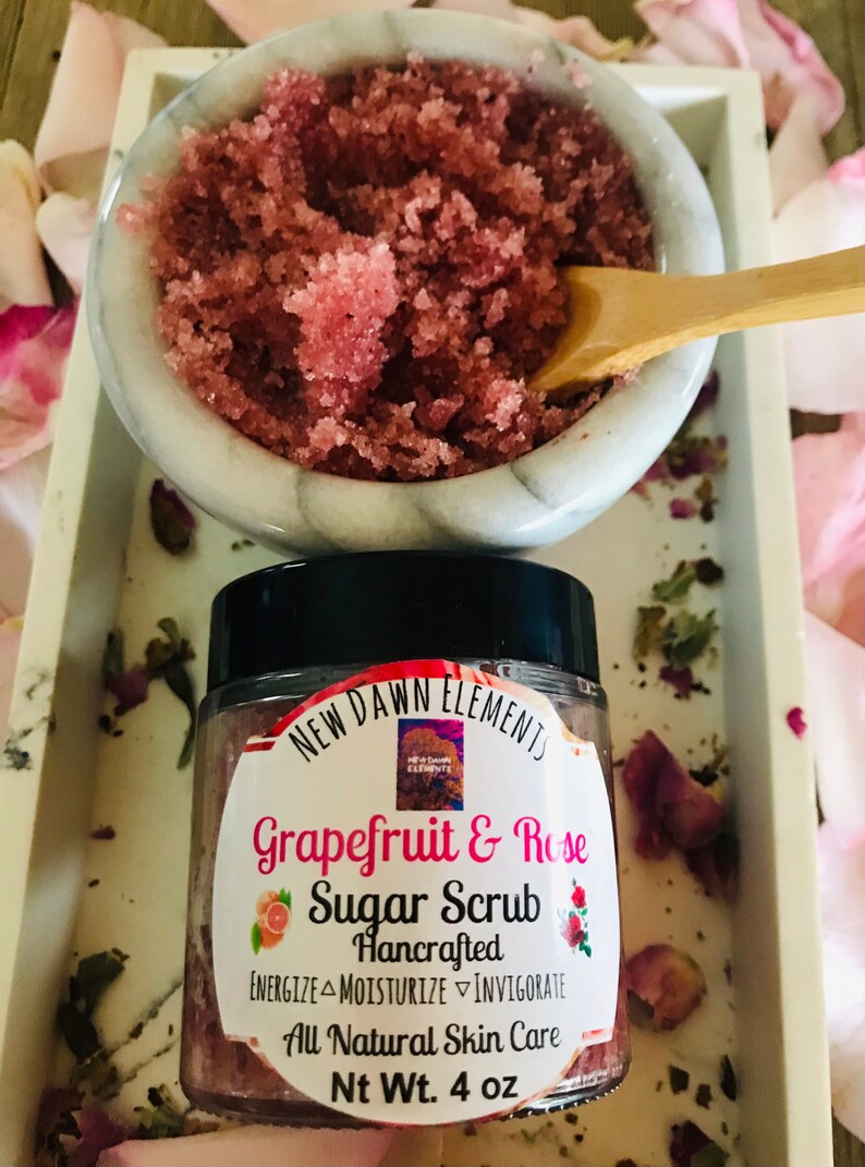 May include: A glass jar of pink grapefruit and rose sugar scrub with a black lid. The label reads "NEW DAWN ELEMENTS Grapefruit & Rose Sugar Scrub Hancrafted ENERGIZE MOISTURIZE INVIGORATE All Natural Skin Care Nt Wt. 4 oz".
