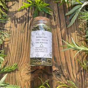 May include: A small glass jar with a cork stopper filled with dried herbs. The label reads "NEW DAWN ELEMENTS CLEANSE + PROTECTION HERBAL INCENSE BLEND". The jar is surrounded by green foliage on a wooden surface, creating a natural, earthy aesthetic.