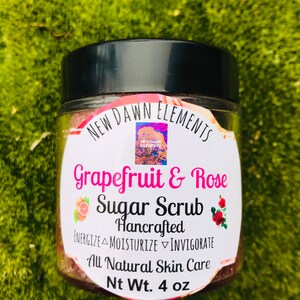 May include: A glass jar with a black lid containing a pink and white sugar scrub. The label reads "NEW DAWN ELEMENTS Grapefruit & Rose Sugar Scrub Hancrafted ENERGIZE MOISTURIZE INVIGORATE All Natural Skin Care Nt Wt. 4 oz".