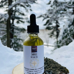 May include: A glass bottle of Imbolc Ritual Oil with a black dropper cap. The bottle is labeled with the words "Imbolc Ritual Oil" and a list of ingredients including cedarwood, basil, and peppermint. The bottle is sitting on a wooden surface covered in snow.