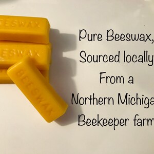 Op de afbeelding: Een stapel van vijf gele bijenwasstaven, elk met het label "BEESWAX". De tekst "Pure Beeswax, Sourced locally From a Northern Michigan Beekeeper farm" staat onder de staven.