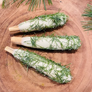 May include: Three white candles wrapped in green pine needles on a wooden surface.