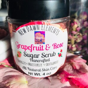 May include: A glass jar with a black lid filled with a pink sugar scrub. The label reads "NEW DAWN ELEMENTS Grapefruit & Rose Sugar Scrub Hancrafted ENERGIZE MOISTURIZE INVIGORATE All Natural Skin Care Nt Wt. 4 oz".