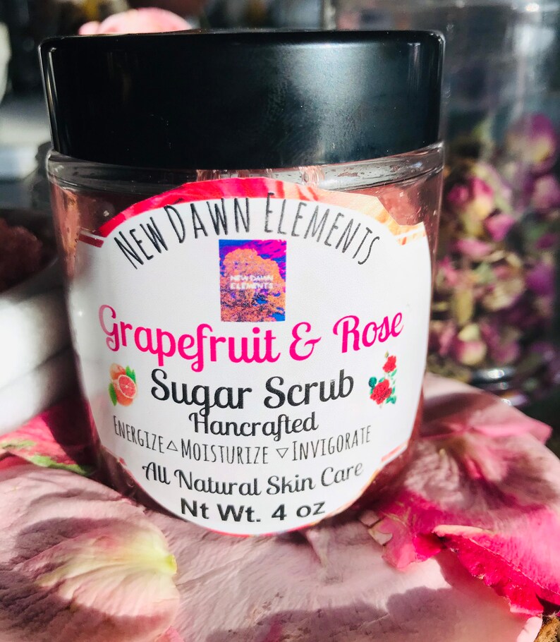 May include: A glass jar with a black lid filled with a pink sugar scrub. The label reads "NEW DAWN ELEMENTS Grapefruit & Rose Sugar Scrub Hancrafted ENERGIZE MOISTURIZE INVIGORATE All Natural Skin Care Nt Wt. 4 oz".