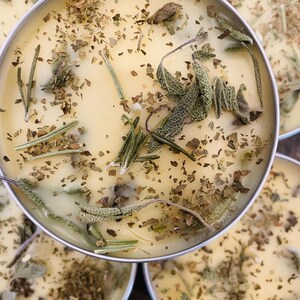 Op de afbeelding: Close-up van vier kleine, ronde, gele kaarsen met gedroogde kruiden en bladeren die in de was zijn ingebed. De kaarsen bevinden zich in metalen containers.