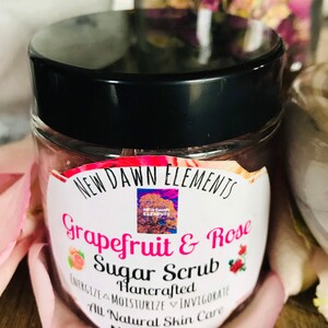May include: A white jar with a black lid filled with a pink sugar scrub. The label reads "NEW DAWN ELEMENTS Grapefruit & Rose Sugar Scrub Hancrafted ENERGIZE MOISTURIZE INVIGORATE All Natural Skin Care Nt Wt. 4 oz".