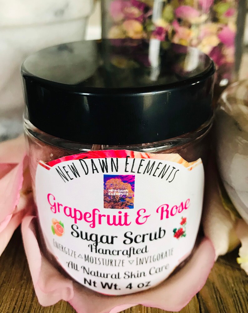 May include: A white jar with a black lid filled with a pink sugar scrub. The label reads "NEW DAWN ELEMENTS Grapefruit & Rose Sugar Scrub Hancrafted ENERGIZE MOISTURIZE INVIGORATE All Natural Skin Care Nt Wt. 4 oz".
