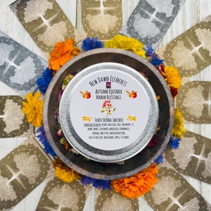 May include: A silver tin of loose herbal incense labeled "New Dawn Elements Autumn Equinox Mabon Blessings Loose Herbal Incense" with a list of ingredients including cedar, mullein, sage, rosemary, St. John's Wort, lavender, marigold, chamomile, yarrow, sweet pea, bachelor button, rose & rose hip, birch bark, apple, & fennel.