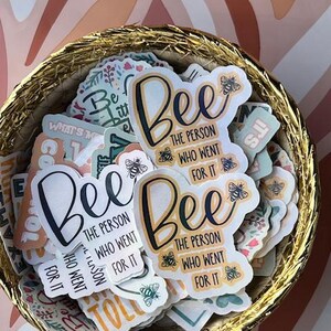 Könnte beinhalten: Ein Korb gefüllt mit verschiedenen Aufklebern mit dem Spruch "Bee the person who went for it" in verschiedenen Farben und Schriftarten. Einige Aufkleber haben eine Bienenillustration.