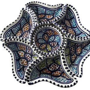 May include: A white ceramic serving dish with a six-sectioned design. The dish features a central circular compartment and six surrounding petal-shaped sections. Each section is decorated with blue, brown, and green patterns, and outlined with a dark blue border.