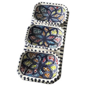 Può includere: Un piatto da portata rettangolare con tre scomparti collegati. Ogni scomparto presenta un colorato motivo floreale in blu, rosso, giallo e verde. Il piatto ha una base bianca con un bordo a righe bianche e nere.