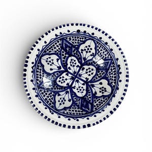 May include: A white ceramic plate with a blue floral design. The plate features a central flower motif surrounded by intricate patterns and a dotted border. The design is hand-painted, showcasing a traditional style.