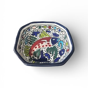 Può includere: Una ciotola in ceramica dipinta a mano con bordo blu scuro e interno bianco. La ciotola presenta un colorato disegno di pesce in rosso, blu e verde, circondato da motivi decorativi. La ciotola ha una forma ottagonale.