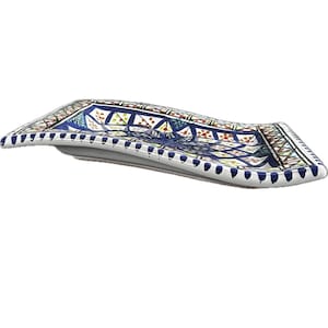 May include: A decorative ceramic serving dish with a curved, rectangular shape. The dish features a detailed, colourful pattern with blue, white, red, and green geometric designs. The edges are trimmed with a dotted pattern.
