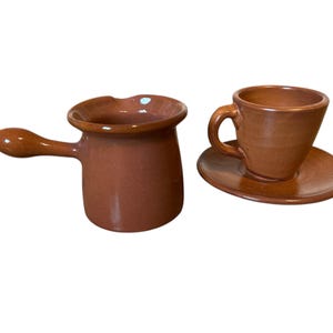 May include: A brown ceramic coffee set, comprising a small pot with a handle and a cup with a saucer. The set has a rustic, handmade appearance. The cup and saucer are small, suitable for espresso.