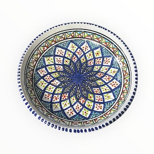 May include: A round, hand-painted ceramic plate with a vibrant floral design. The plate features a deep blue central flower, surrounded by intricate patterns in white, green, red, and blue. The rim is decorated with small blue dots.