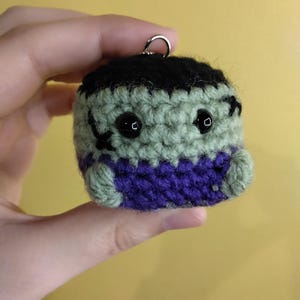 May include: A small, round, crocheted plush keychain featuring a light green face with black yarn hair, two black bead eyes, and black stitched scars. The figure wears a purple crocheted shirt and has small light green arms.