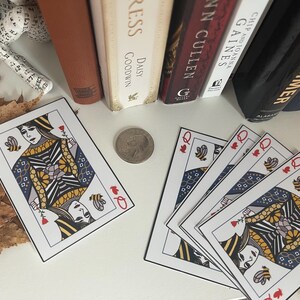 Puede incluir: Cartas de juego con el dise&ntilde;o de la Reina de las Abejas, con detalles de abejas y flores. Las cartas est&aacute;n dispuestas sobre una superficie blanca con una moneda y libros en el fondo. Las cartas tienen un borde blanco y un s&iacute;mbolo de palo rojo.