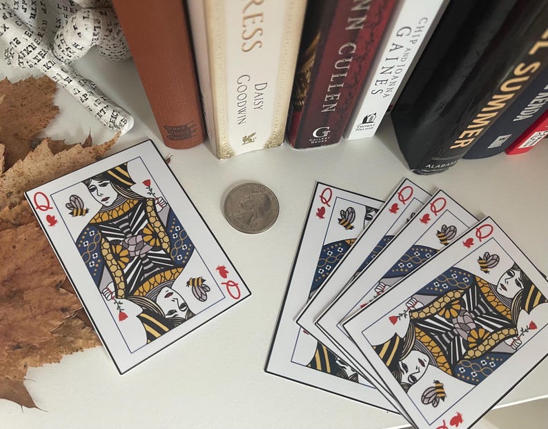 Puede incluir: Cartas de juego con el dise&ntilde;o de la Reina de las Abejas, con detalles de abejas y flores. Las cartas est&aacute;n dispuestas sobre una superficie blanca con una moneda y libros en el fondo. Las cartas tienen un borde blanco y un s&iacute;mbolo de palo rojo.