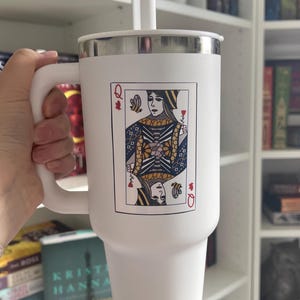 Puede incluir: Vaso t&eacute;rmico blanco con borde y asa plateados. El vaso presenta un dise&ntilde;o de carta de la Reina de Corazones. Incluye pajita y tapa blancas. Perfecto para bebidas fr&iacute;as o calientes.
