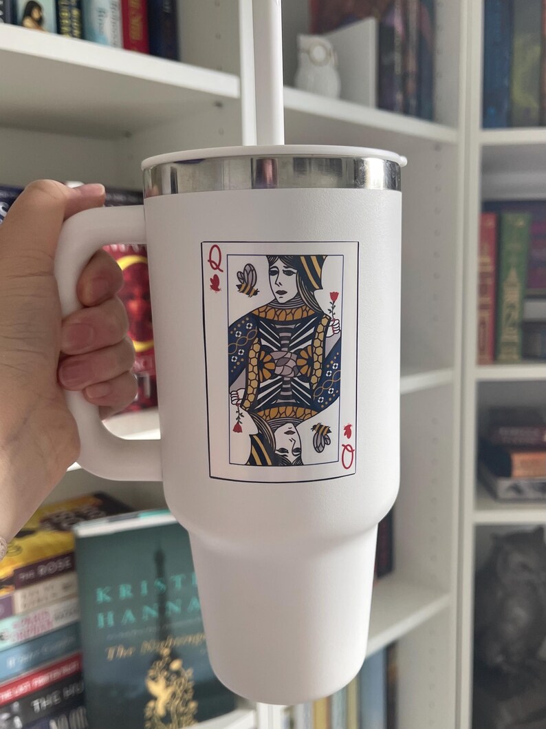 Puede incluir: Vaso t&eacute;rmico blanco con borde y asa plateados. El vaso presenta un dise&ntilde;o de carta de la Reina de Corazones. Incluye pajita y tapa blancas. Perfecto para bebidas fr&iacute;as o calientes.