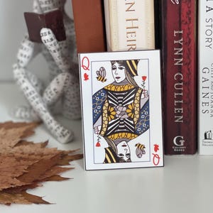 Puede incluir: Una carta de la Reina de Corazones, con un s&iacute;mbolo de coraz&oacute;n rojo y la letra "Q". La carta presenta una ilustraci&oacute;n detallada de una reina sosteniendo una rosa, con una abeja y un dise&ntilde;o floral. La carta est&aacute; colocada frente a libros.