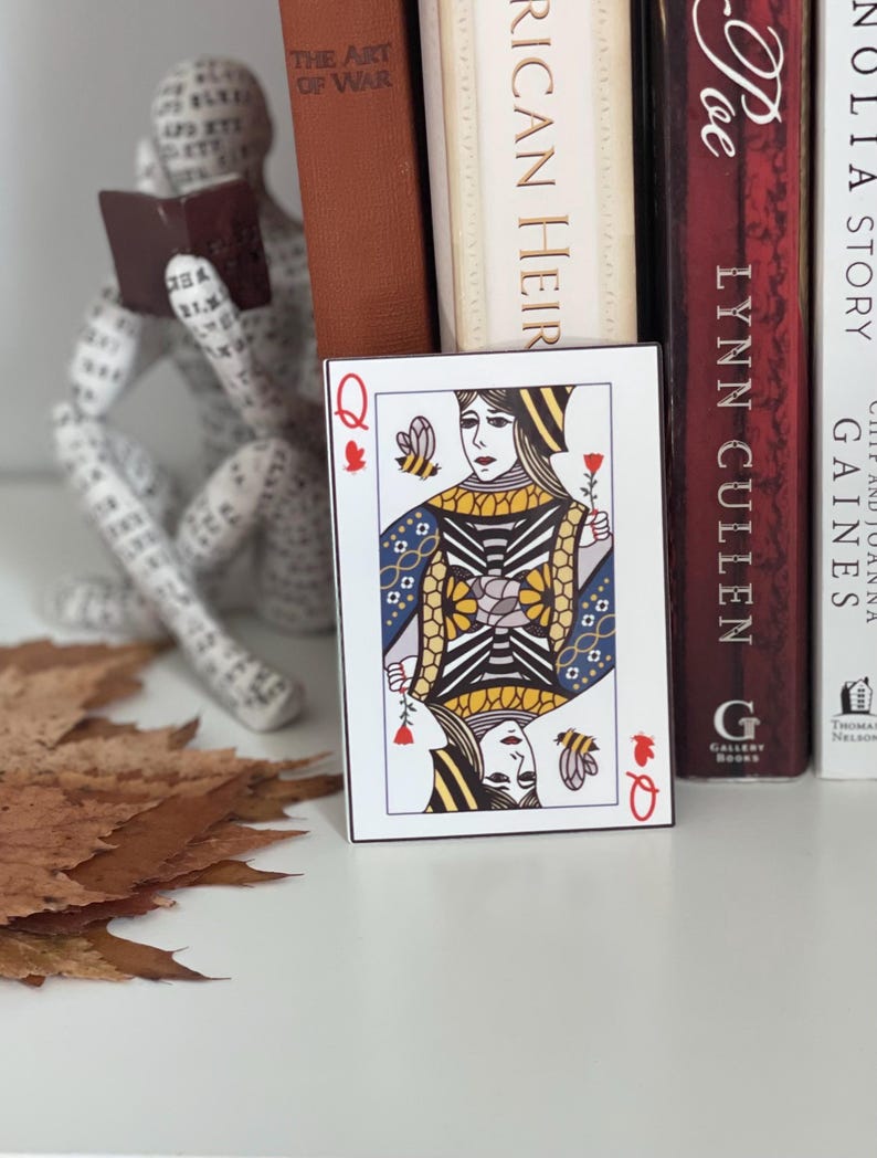 Puede incluir: Una carta de la Reina de Corazones, con un s&iacute;mbolo de coraz&oacute;n rojo y la letra "Q". La carta presenta una ilustraci&oacute;n detallada de una reina sosteniendo una rosa, con una abeja y un dise&ntilde;o floral. La carta est&aacute; colocada frente a libros.