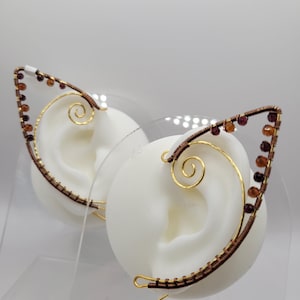 May include: A pair of handcrafted ear cuffs designed to resemble cat ears. The ear cuffs are made with copper wire, adorned with small, reddish-brown beads, and feature a gold-colored swirl design. The ear cuffs are displayed on white ear-shaped forms.