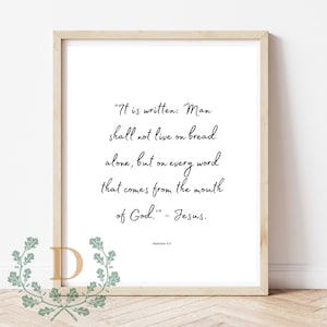 Op de afbeelding: Een printbaar wanddecor ontwerp met een zwart-wit typografie citaat uit de Bijbel: "Er staat geschreven: De mens zal niet van brood alleen leven, maar van elk woord dat uit de mond van God komt." - Jezus. Mattheüs 4:4