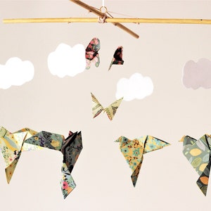 May include: A mobile with origami birds and butterflies, featuring a floral pattern in shades of yellow, green, and pink. The mobile is suspended from a wooden rod and includes white and purple clouds.