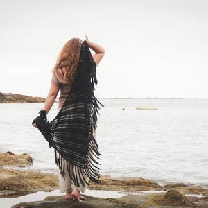 以下が含まれることがあります： フリンジの付いた黒いショールを羽織った女性が、岩だらけのビーチに立って海を見つめている