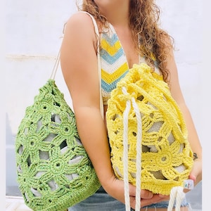 Puede incluir: Dos bolsas de cordón de ganchillo, una verde y otra amarilla, con asas de cuerda blanca. Las bolsas están hechas con un patrón de flores.