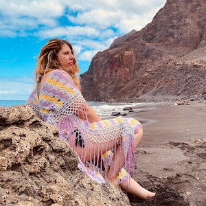 Puede incluir: Un encubrimiento de ganchillo en tonos lavanda, amarillo y blanco con detalles de flecos. El encubrimiento se usa en una playa rocosa con una montaña al fondo. El cielo es azul con nubes blancas.