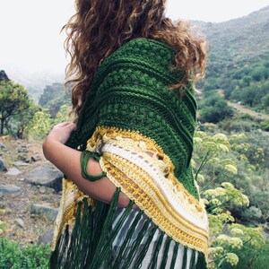 Puede incluir: Un chal de ganchillo verde, amarillo y blanco con flecos. El chal está drapeado sobre los hombros. El diseño del chal presenta un patrón texturizado en verde, que transiciona a amarillo y blanco con un borde con flecos. El fondo muestra un entorno natural al aire libre.