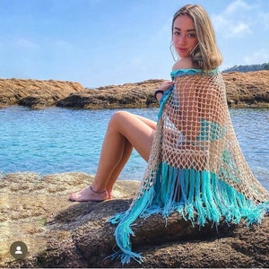 Puede incluir: Un chal de crochet beige y turquesa con flecos. El chal está drapeado sobre los hombros. El fondo muestra una costa rocosa y el océano.