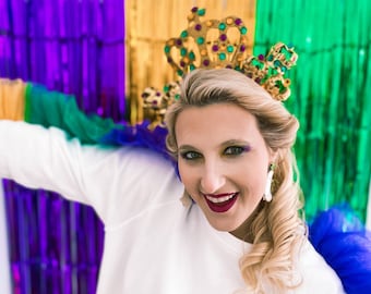 May include: A woman wearing a gold crown with green and purple jewels, a white shirt, and a blue and green scarf. She is smiling and looking at the camera.