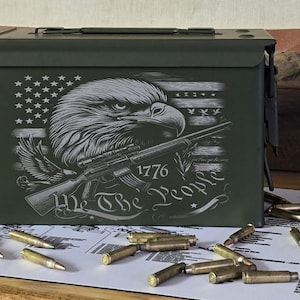 May include: A green metal ammunition box with a graphic of an eagle, rifle, and American flag. The text "We The People" and "1776" are also displayed. Several bullets are scattered around the box.