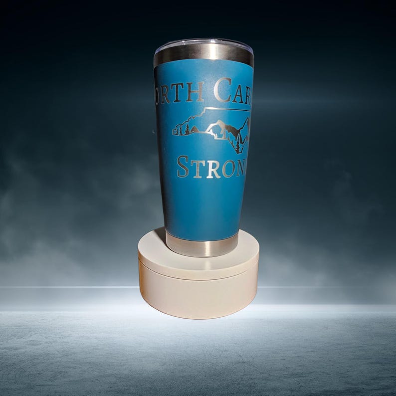 May include: A blue stainless steel tumbler with a silver outline of the state of North Carolina and the words "North Carolina Strong" engraved on the side.
