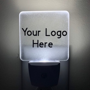 May include: A square, frosted, illuminated night light plugged into a wall outlet. The front surface features the black text 'Your Logo Here' centered on the translucent panel, which emits a soft white glow from the base unit below it.
