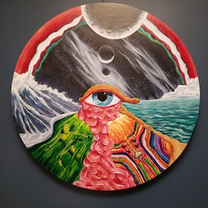 May include: A circular painting with a surreal and abstract design. The painting features a large, pink, fleshy eye with a blue iris, surrounded by a swirling, colorful background. The background includes a green landscape, a blue ocean, and a dark sky with white clouds and three gray planets.