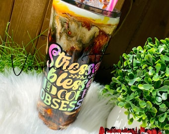 May include: A stainless steel tumbler with a brown and white marble design. The tumbler has a pink straw and is filled with ice and a brown liquid. The tumbler has a rainbow iridescent sticker with the text "Stressed Blessed and Coffee Obsessed".