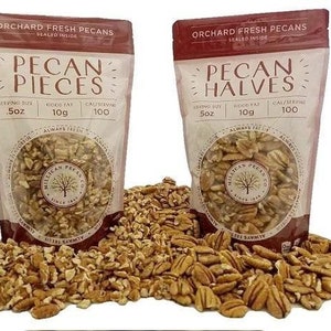 May include: Two bags of pecan pieces and pecan halves from the Milligan Pecan Co. The bags are white with brown text and a brown logo. The bags are sitting on a pile of pecans.