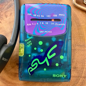 May include: A vintage Sony SRF-PSY03 portable radio with a teal and purple design. The radio features an FM/AM tuner, power switch, and the text "SONY". The face has a purple section with the frequency range and the text "SRF-PSY03".