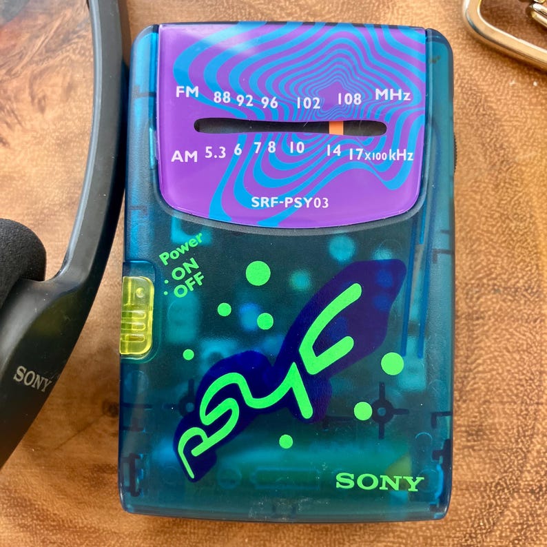 May include: A vintage Sony SRF-PSY03 portable radio with a teal and purple design. The radio features an FM/AM tuner, power switch, and the text "SONY". The face has a purple section with the frequency range and the text "SRF-PSY03".