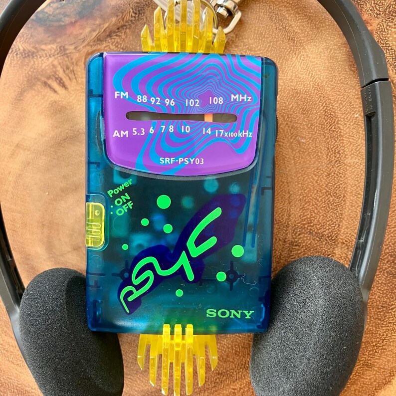 May include: A vintage blue and purple Sony SRF-PSY03 radio with a retro design. The radio features FM and AM frequency dials, a power switch, and the word "SONY". The radio is paired with black headphones and a yellow plastic keychain.