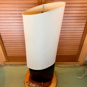 May include: A tall, modern lamp with a white, angled shade and a black base. The lamp sits on a round, wooden base. The shade is off-white and the base is black. The lamp is plugged in and ready to use.