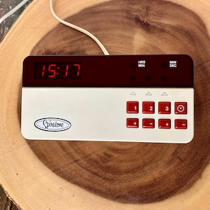 May include: A rectangular Spiratone digital timer with a white base and a dark display panel showing red 15:17 digits. The device features a grid of red buttons labeled with numbers 1, 2, 3, plus and minus signs, and a clock icon, with a white power cord.